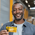 Smiling certified forklift operator proudly holding up his certification card inside a warehouse